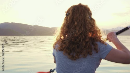 Happy young woman with her dog Jack Russell Terrier paddling on an inflatable kayak on the water of a large mountain lake against a beautiful orange sunset. Family Sports Weekend
