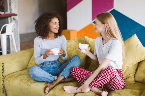 Beautiful african american girl with dark curly hair and pretty girl with blond hair sitting on sofa holding cups of coffee in hands while happily spending time together at home