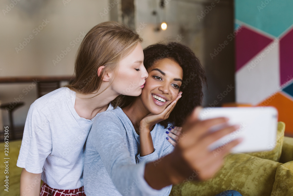 Young attractive woman with dark curly hair happily taking photos on cellphone while pretty woman with blond hair kissing her in cheek spending time together at cozy home Young attractive woman with dark curly hair happily taking photos on cellphone while pretty woman with blond hair kissing her in cheek spending time together at cozy home