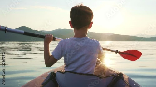 Slow motion of a happy boy with his dog Jack Russell Terrier paddling an inflatable kayak on the water of a large mountain lake against the backdrop of beautiful orange sunset. Family sports vacation