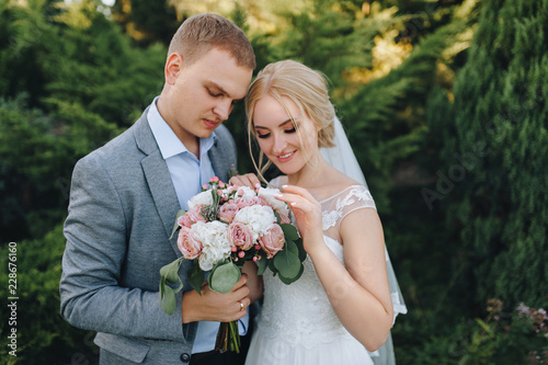 Beautiful newlyweds are considering a bouquet in a green flowered garden, in nature and smiling. Wedding portrait of a stylish groom and young bride. Wedding photography. photo