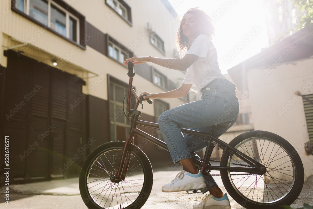 Beautiful african american girl with dark curly hair in white T-shirt and jeans dreamily looking in camera while riding bicycle outdoor