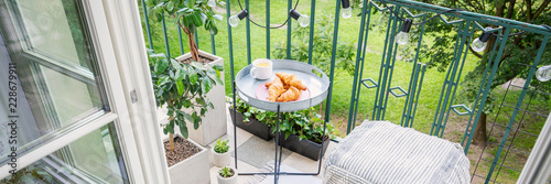 Croissants and tea on the table on bright balcony with pouf and green plants in pots, panoramic view, real photo
