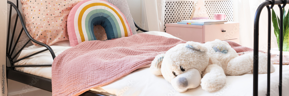Close-up of child bed with teddy bear, pastel pink blanket and rainbow cushion in the real photo - obrazy, fototapety, plakaty 