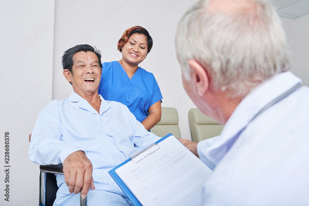 Anonymous elderly doctor and cheerful nurse looking at laughing senior man on wheelchair in hospital Anonymous elderly doctor and cheerful nurse looking at laughing senior man on wheelchair in hospital