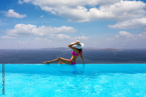 Attraktive Frau im Bikini und mit weißem Hut genießt die Aussicht von einem Pool auf das Meer 