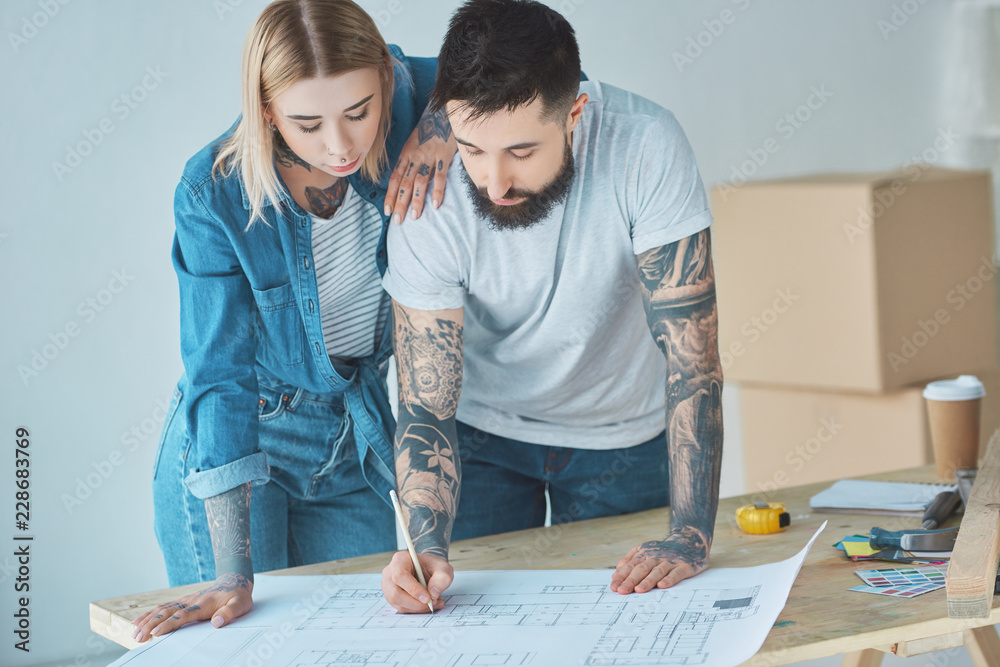 portrait of tattooed couple looking at blueprints on wooden tabletop at new home portrait of tattooed couple looking at blueprints on wooden tabletop at new home