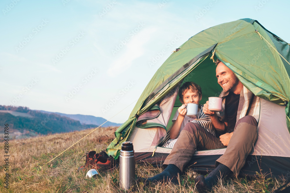 Family lisure concept image. Father and son drink a tea sitting in touristic tent - obrazy, fototapety, plakaty 