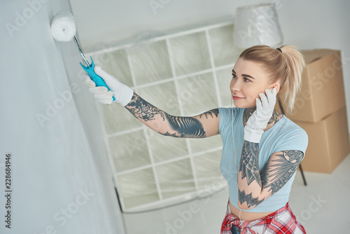 side view of young tattooed woman in earphones painting wall at new home