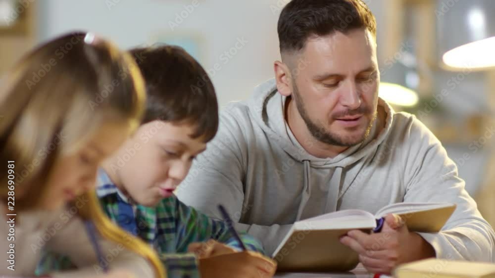 Close-up of diligent kids doing homework together with father: brother and sister writing down in notepads while dad reading textbook aloud Close-up of diligent kids doing homework together with father: brother and sister writing down in notepads while dad reading textbook aloud