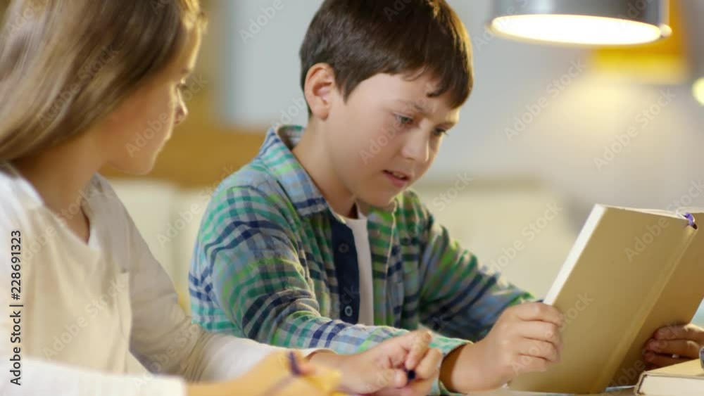 Rack focused shot of diligent kids doing homework together: cute girl writing down in notepad while brother reading textbook aloud Rack focused shot of diligent kids doing homework together: cute girl writing down in notepad while brother reading textbook aloud