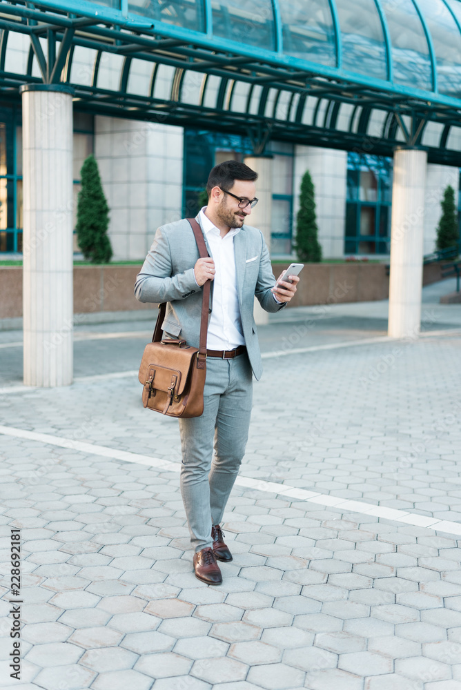 Young successful happy businessman looking at the phone and smiling. Wearing elegant suit, business bag and modern glasses.