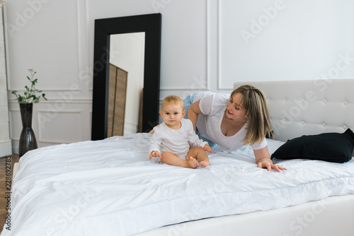 Beautiful happy mother and baby smiling and playing on the bed in the morning wearing pajamas while mommy is looking at the baby and baby is looking at camera.