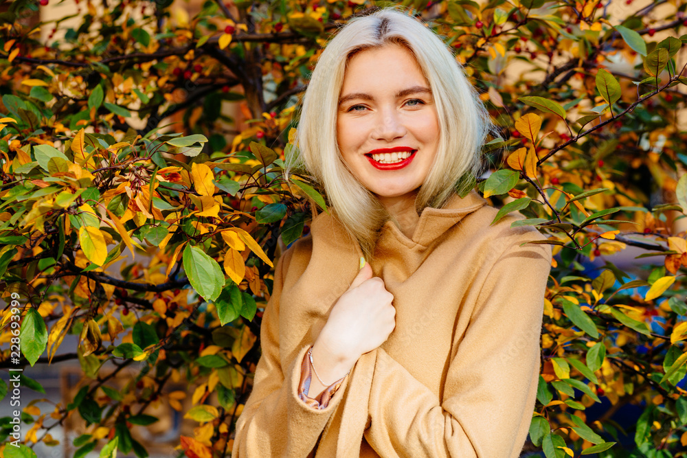 Close up outdoor portrait of young blond happy smiling woman with lips wearing flesh-coloured coat posing near autumn tree. Model with red lips, long blond hair. Lady looking at camera