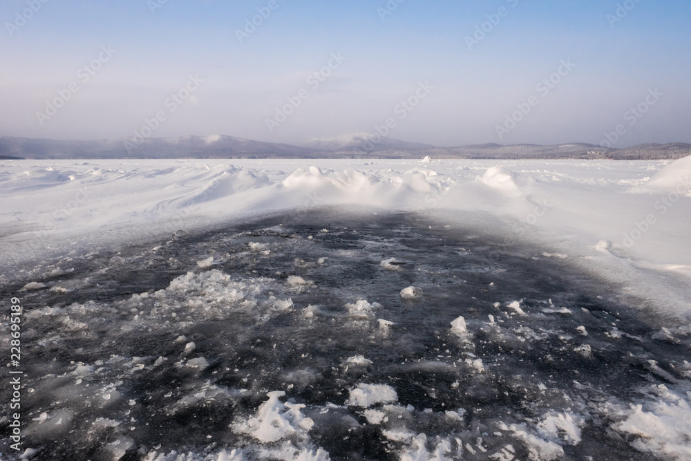 Obraz premium Natural winter lake ice breaking with clear sky background, selective focus. Frozen mountain lake with blue ice and cracks on the surface. Winter landscape with snowy hills under a blue sky.