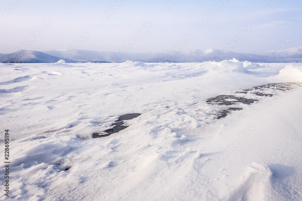 Natural winter lake ice breaking with clear sky background, selective focus. Frozen mountain lake with blue ice and cracks on the surface. Winter landscape with snowy hills under a blue sky. Natural winter lake ice breaking with clear sky background, selective focus. Frozen mountain lake with blue ice and cracks on the surface. Winter landscape with snowy hills under a blue sky.