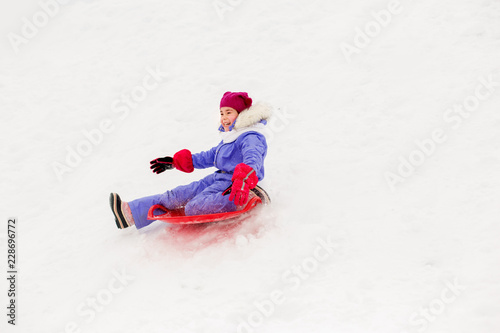 childhood, sledging and season concept - happy little girl sliding down on snow saucer sled outdoors in winter