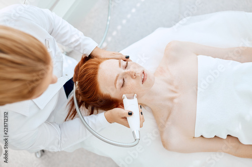 Apparatus face treatment in medical spa center, beautiful redhead lady receiving electric facial peeling massage. Cosmetological methods, apparatus cosmetology, SPA-procedures Concept.