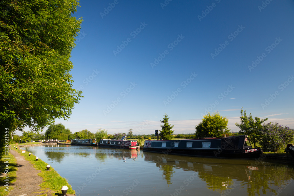 Hertfordshire, UK. Narrowboats moored along a wide canal under blue skies on a summer's morning.