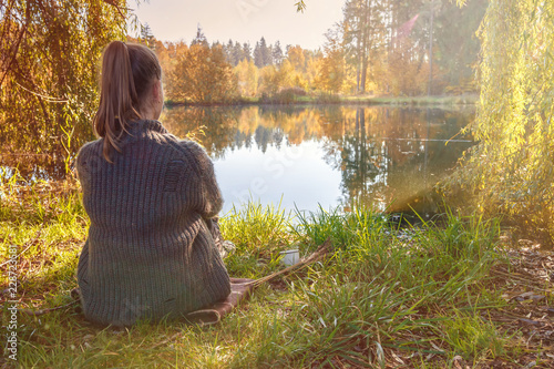 young girl is sitting on the shore of the lake on a warm cozy autumn afternoon, lifestyle is authentic.