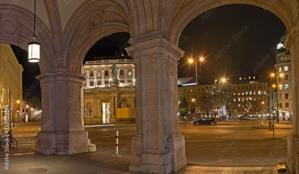 View of illuminated Albertina square at night through arches on pillars of the state opera in Vienna.
