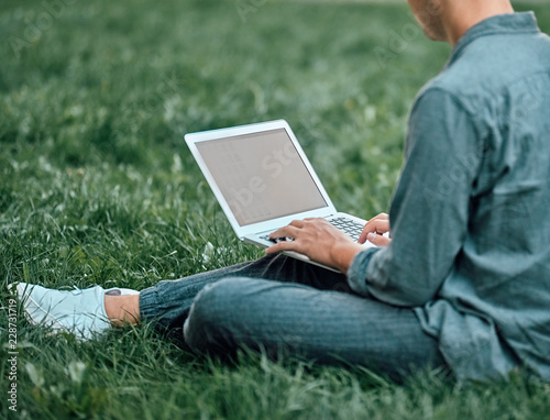 A guy is sitting on the grass and looking at the laptop