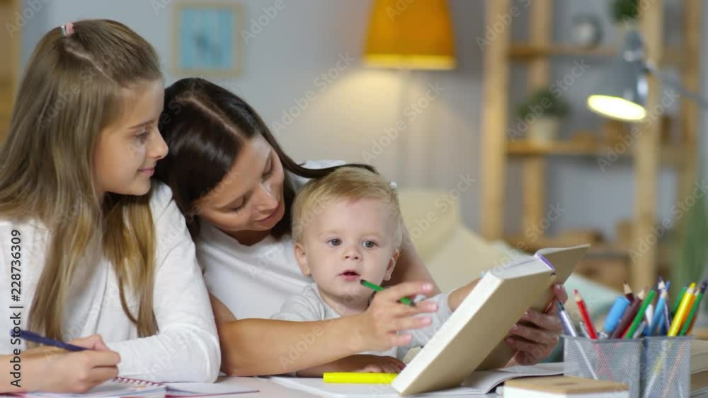 Young mother reading textbook and explaining daughter how to do homework while cute baby boy sitting on her knees and playing with pencil Young mother reading textbook and explaining daughter how to do homework while cute baby boy sitting on her knees and playing with pencil
