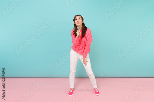 Full length portrait fun woman in knitted sweater, white pants, headphones sing song in microphone isolated on bright pink blue pastel wall background in studio. Lifestyle concept. Mock up copy space.