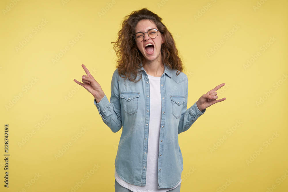 Young brunette woman with curly hair shows crazy expression doing rock symbol with hands up Young brunette woman with curly hair shows crazy expression doing rock symbol with hands up