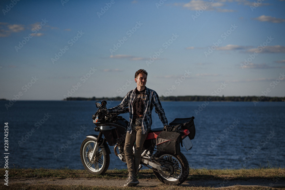 Obraz premium Relaxed motorcyclist stands with his motorbike beside the natural lake. Near the lake.