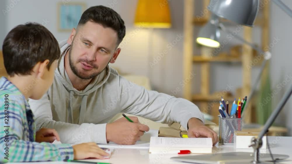 Dad sitting with son at desk in home, reading textbook and discussing something while doing homework together Dad sitting with son at desk in home, reading textbook and discussing something while doing homework together