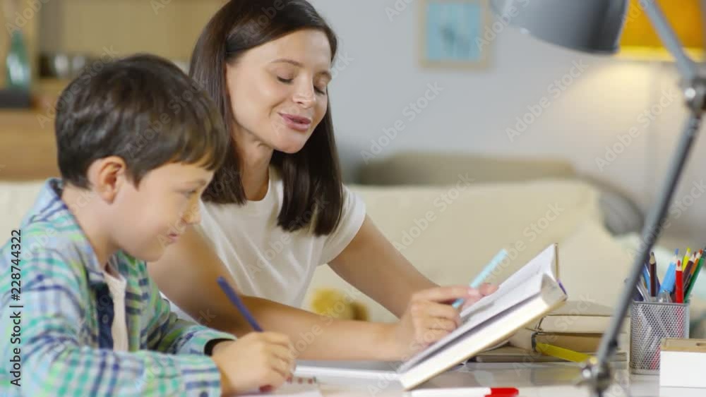 Beautiful mother reading textbook and explaining something to son while doing homework together in the living room Beautiful mother reading textbook and explaining something to son while doing homework together in the living room