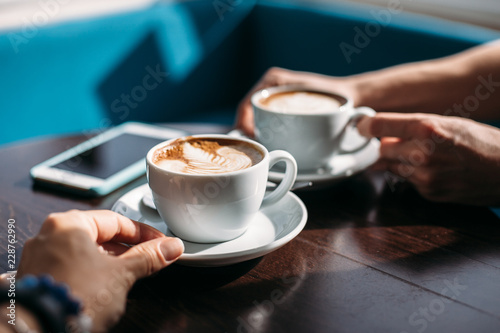 Two cups of cappuccino with latte art on wooden table in the hands of man and woman.