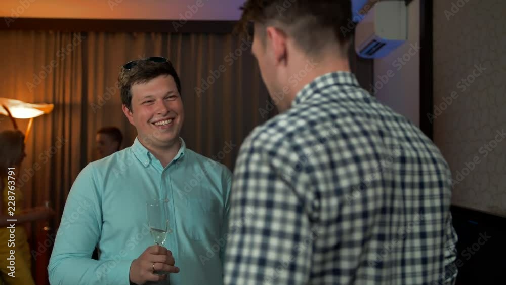 Two smiling guys speaking at private home flat apartment party. Their friends also have conversation at background. Sparkling wine champigne welcome drinks. Happy smiling faces.