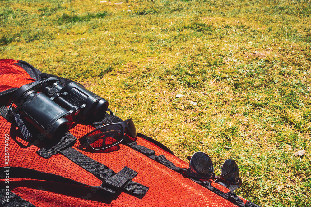 Big red camping backpack with traveler items (sunglasses, binocular) on grass in sunny day. Active rest. Hiking on nature. Mountain trekking. Amazing leisure activity in highlands.