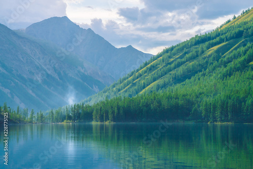 Fototapeta Naklejka Na Ścianę i Meble -  Ghostly mountain lake in highlands at early morning. Beautiful misty mountains reflected in calm clear water surface. Smoke of campfires. Amazing atmospheric foggy landscape of majestic nature.