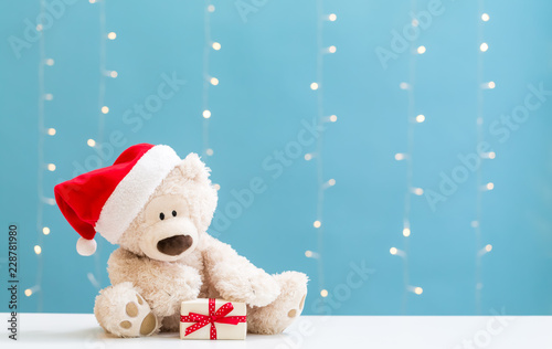 Teddy bear wearing a Santa hat and a gift box on a shiny light blue background