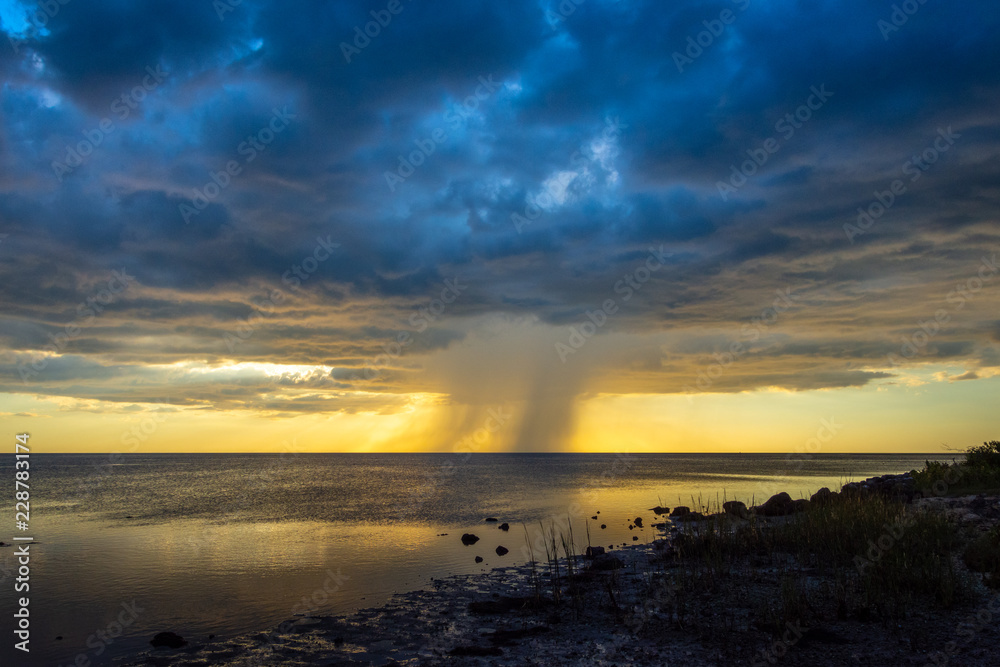 A small rain squall moves out to sea over the Gulf of Mexico after briefly falling on shore, as the sun sets behind it, creating a beautiful and haunting scene..
