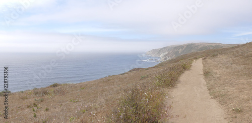 Panorama of a dirt walking track atop a seaside cliff with mist over the water