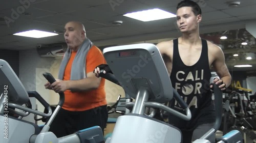Fat funny man eating hamburger, training on an ellipsoid, near him young handsome sportsman with a nice fitness body actively trains on another ellipsoid.