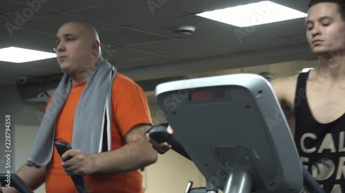 Fat funny man with a towel on his shoulders training on an ellipsoid and eating hamburger, near him young handsome sportsman  actively trains on another ellipsoid. Camera moving