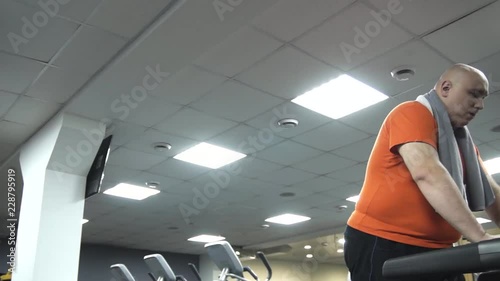Fat man walking on the treadmill, wiping his head with a towel. Camera moving from left to the right. The view from the bottom up