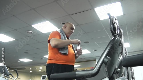 Fat man coming up to the treadmill, drinking water, putting some bottoms and starting to walk on the treadmill. The view from the bottom up