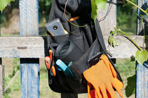 A tool belt bag with acoustic column and orange gloves hanging on a wooden fence with grape leaves