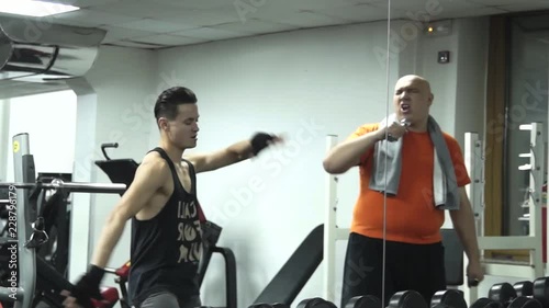 Fatty funny man and a young handsome sportsman are dancing in a gym near dumbells acting a fool