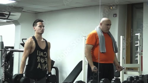 Fatty man picks up the smallest dumbbells standing next to a handsome athlete picking up big dumbbells.Both are trying and tired