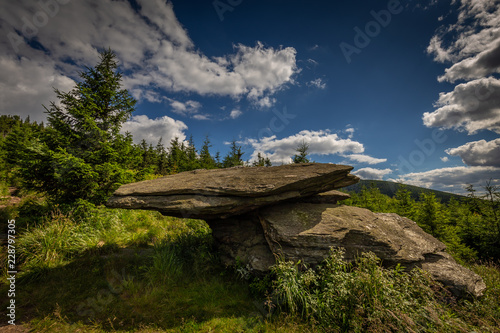 Big flat rock sitting on other on Obri Skaly with green grass, pine trees and dark blue cloudy sky under hill Serak in Jeseniky, Czech Republic photo