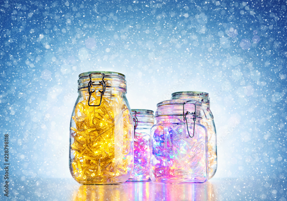 Glass jars with colored luminous garlands inside, on bright blue background with snow. Festive mood concept Glass jars with colored luminous garlands inside, on bright blue background with snow. Festive mood concept