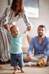 Smiling young family playing and baby learning to walk at home.
