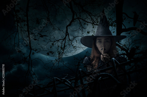 Halloween witch showing silence sign with finger over lips standing over dead tree, crow, birds, full moon and spooky cloudy sky, Halloween mystery concept